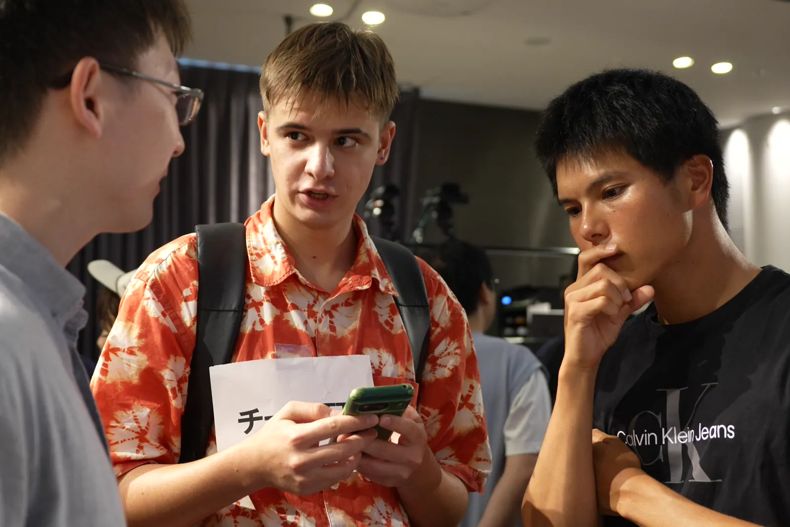 スマートフォンと紙を持って話す男性とそれを聞く2人