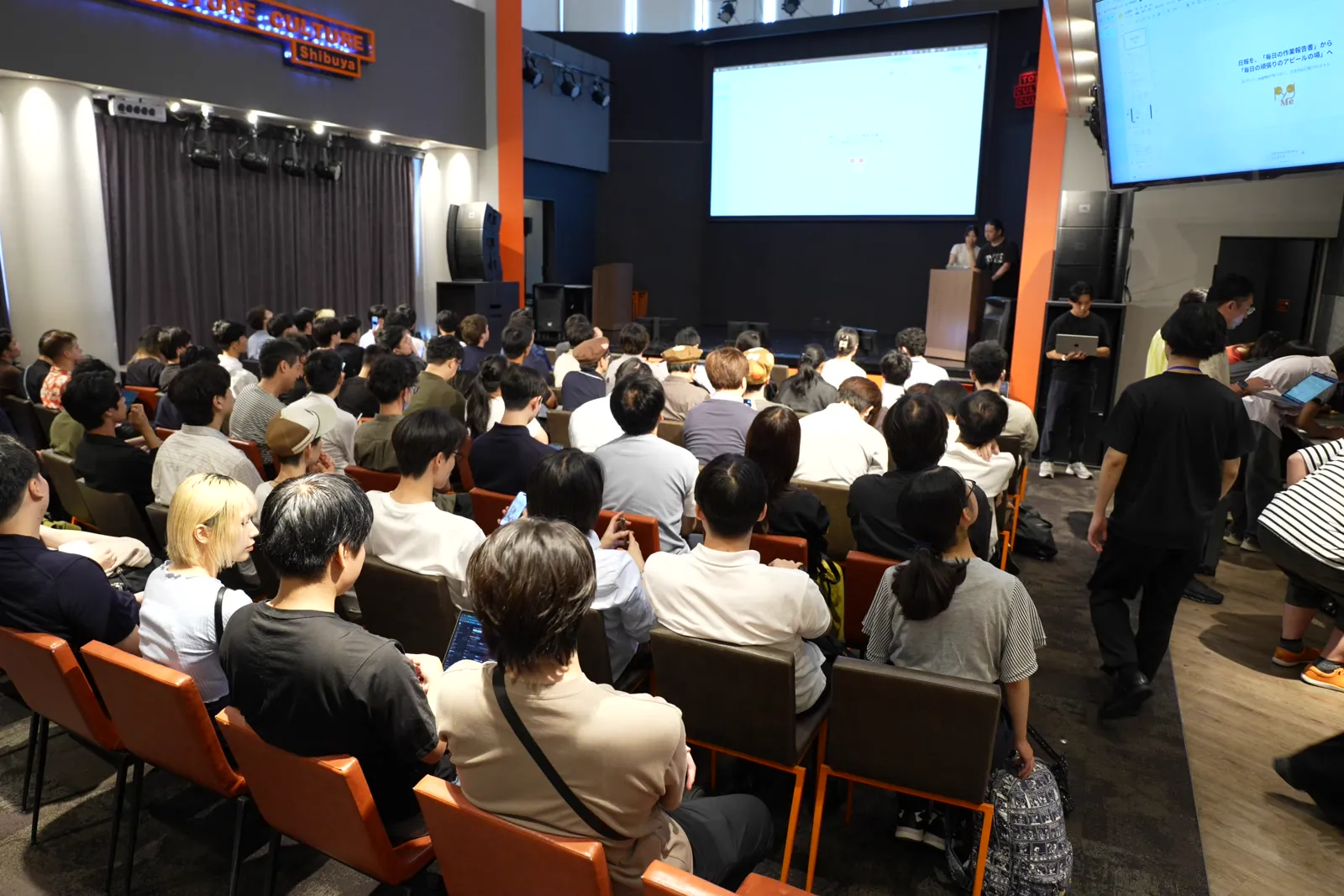 客席が埋まったホールでスクリーンの発表を見る参加者たち